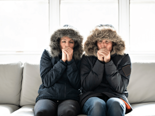couple sitting on couch in living room wearing winter jackets and trying to warm up hands because heater is broken